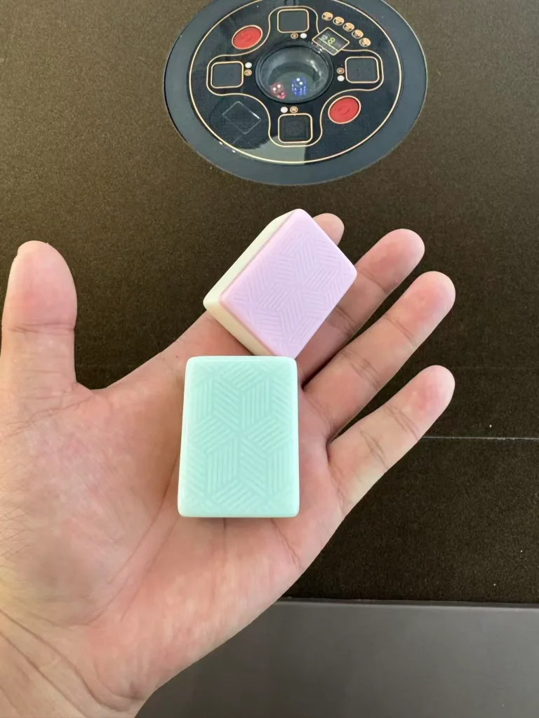 浅绿与浅粉两颗麻将牌静卧掌心，表面纹路细腻，色泽柔和（Two pastel mahjong tiles — mint green and soft pink — resting in an open palm, showing fine engraved textures.）